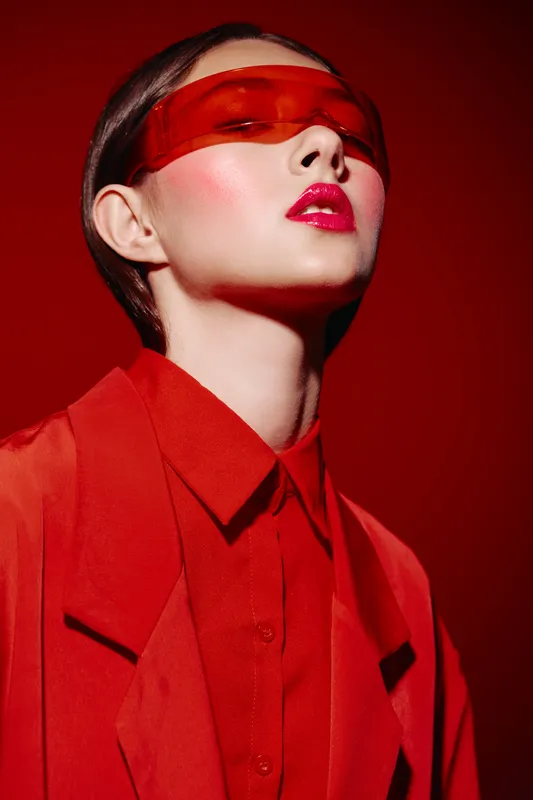 Young woman in vibrant red outfit and translucent orange visor poses confidently against red background. Creative fashion and beauty concept focused on bold color.