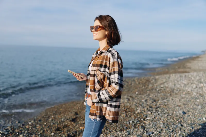 Woman on a beach in a plaid shirt and jeans, holding a smartphone, wearing sunglasses, with calm sea and clear sky behind her, casual lifestyle portrait by the shore