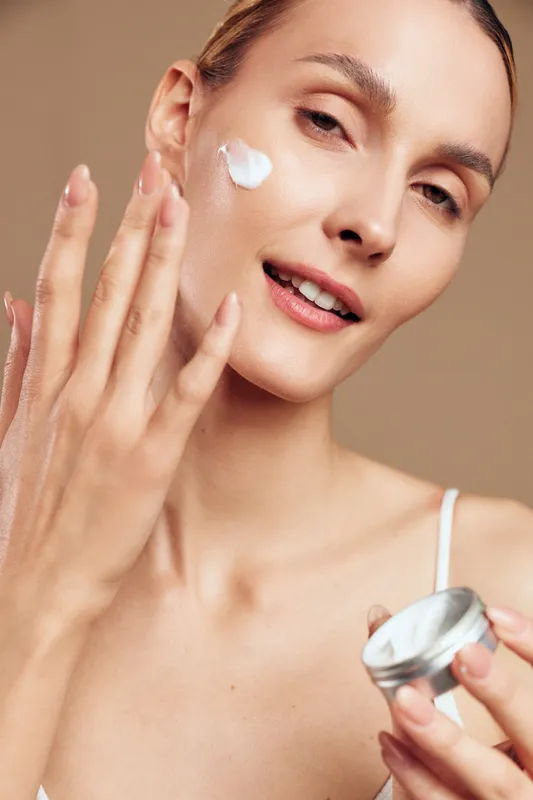A young woman with clear skin applies white facial cream on her cheek using fingers. The close-up shot highlights skincare, beauty routine, and self-care concepts.