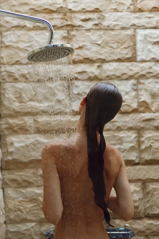 woman showering outdoors under rain showerhead with wet long hair and natural stone wall background, wellness and relaxation moment