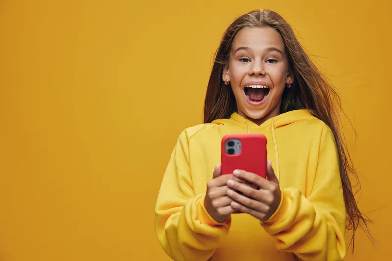 Girl in yellow hoodie holding red smartphone with excited expression looking at camera on yellow background. Young female teenager smiling with surprise and joy while using mobile device indoors.