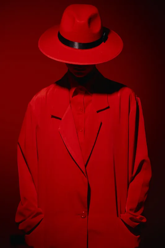 Stylish model wearing a wide-brimmed hat and oversized jacket hides face under red lighting core texture creating bold fashion and beauty creative theme with mystery.
