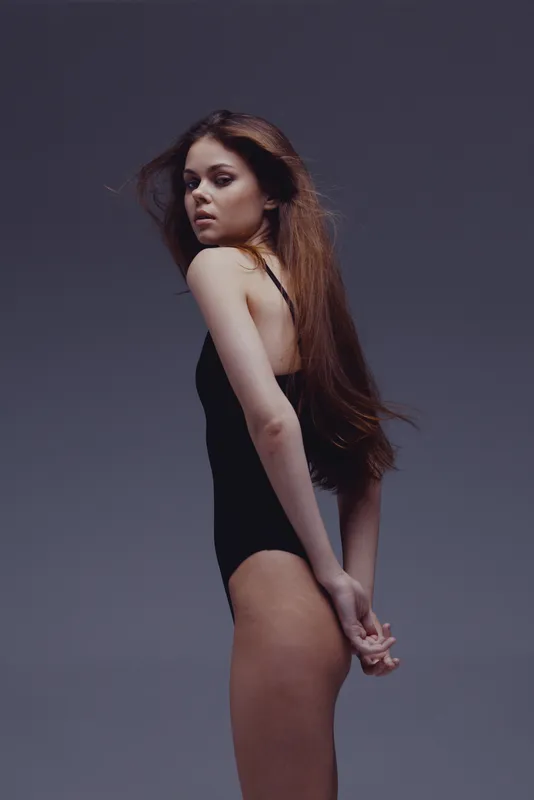 Young woman in black bodysuit posing against a neutral gray background with serious expression and long hair