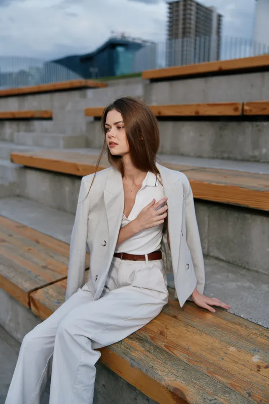 Stylish businesswoman in light attire appearing thoughtful against a modern architectural backdrop, showcasing elegance and professionalism in an urban setting.