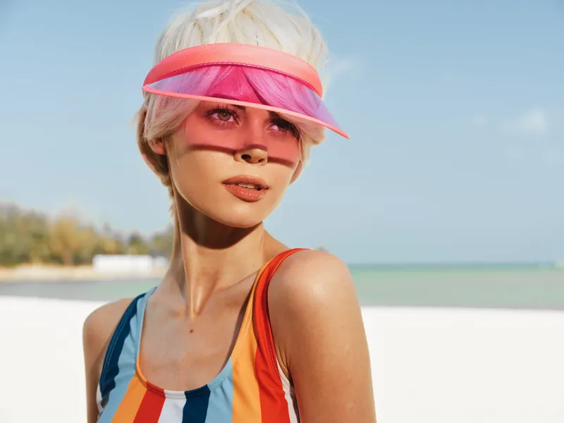 A stylish beach portrait featuring a sunlit woman in a colorful striped swimsuit and pink visor, capturing carefree summer style and vibrant coastal glow.