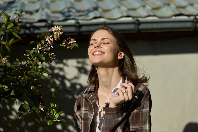 A cheerful woman enjoys a sunlit moment in a casual outdoor setting, eyes closed, face tilted toward the warmth. A lifestyle moment capturing ease and happiness