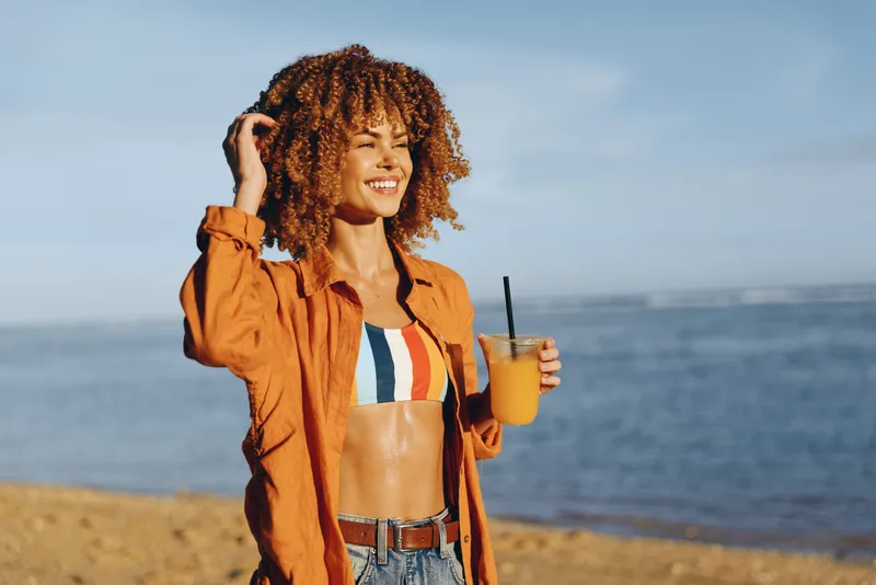 Woman at beach, smiling in sunlight, curly hair blowing, hand holding hair, holding drink, orange jacket and denim, seaside background, summer vacation mood, with blue ocean horizon
