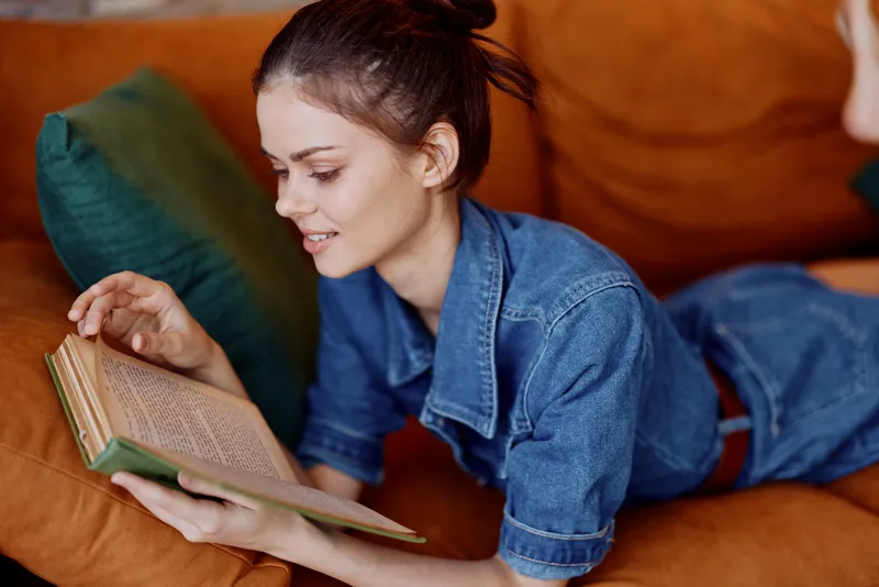 Женщина отдыхает на диване дома, читает книгу, одета в джинсовую рубашку
