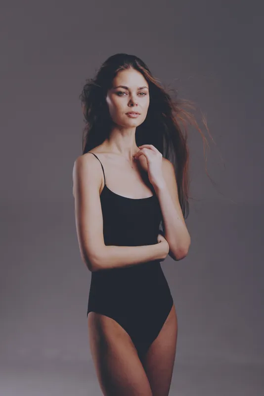 Young woman in black bodysuit posing with serious expression against gray background in soft natural light.