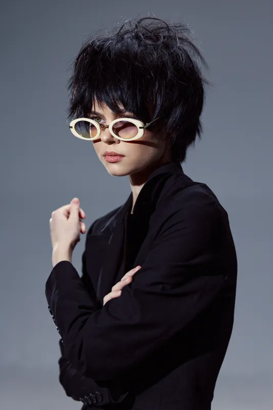 Stylish woman with short black hair wearing white glasses and a black outfit posing confidently in a studio with a gray background.