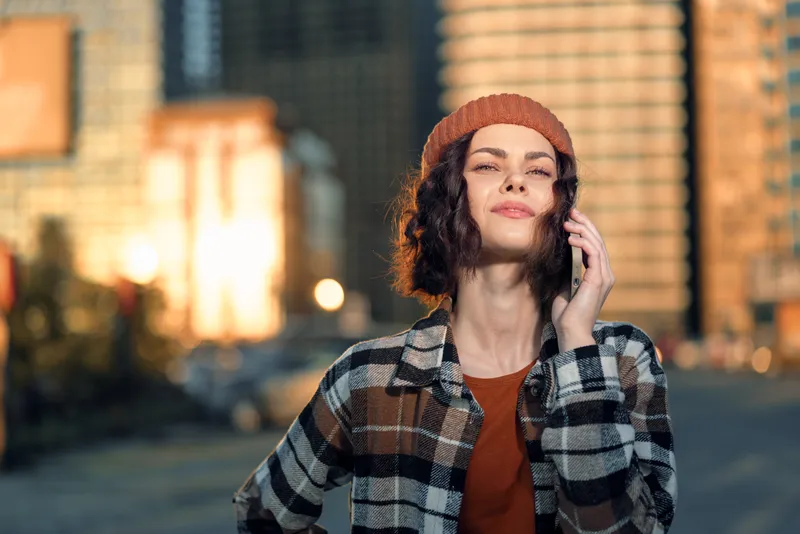 Woman on phone in urban street at golden hour, beanie and plaid shirt, confident smile and candid lifestyle portrait conveying authenticity, mindful living and emotional storytelling.