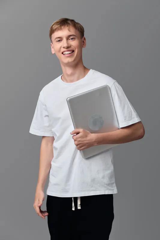 Young man with a laptop, casual attire, smiling, in a studio setting, highlighting technology, education, and modern lifestyle, emphasizing a digital device and youth energy