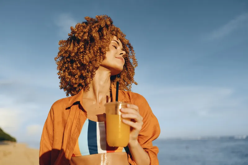 Young woman with curly hair wearing an orange shirt on a sunny beach drinks orange juice through a straw, basking in warm light and a relaxed, carefree mood.