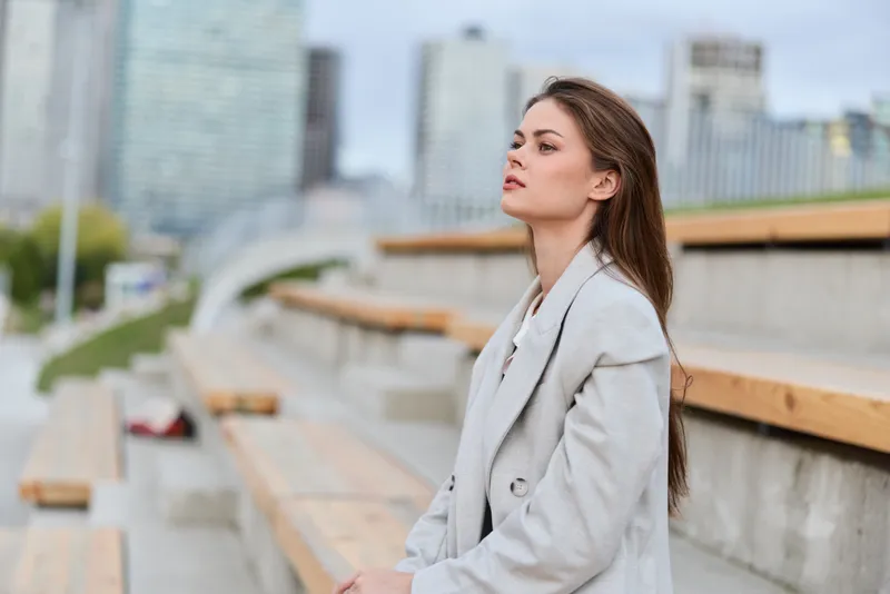 Woman in light coat sitting outdoors on urban wooden bleachers contemplating with cityscape background, autumn casual fashion and pensive mood