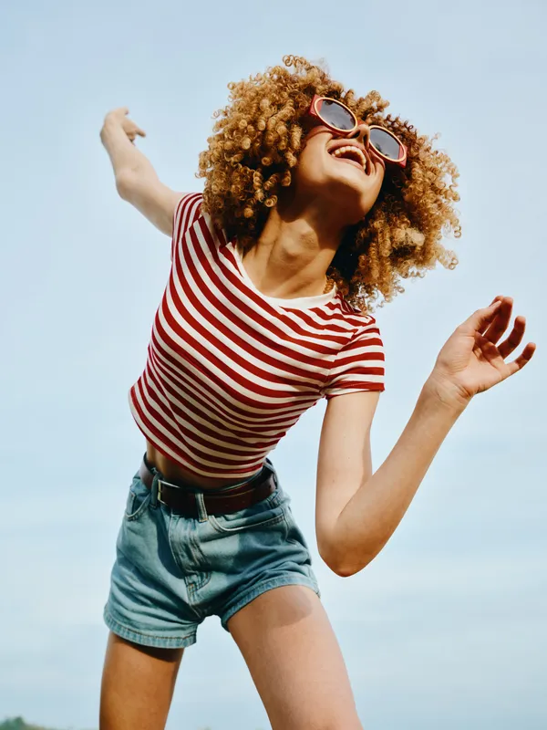 Bright joyful portrait of a young woman with curly hair, sunglasses, striped tee, and denim shorts, jumping with carefree energy against a clear blue sky outdoors.