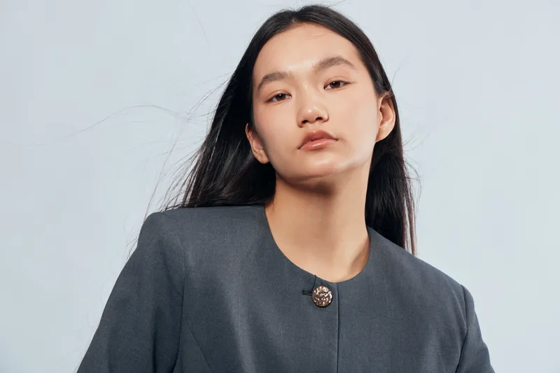 Portrait of confident young woman with long black hair wearing dark jacket standing against light background for fashion, style, beauty, and modern look in studio environment.