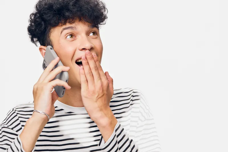 Surprised young man talking on smartphone, wearing striped shirt with expressive emotions and curly hair, isolated on white background, casual lifestyle moment.