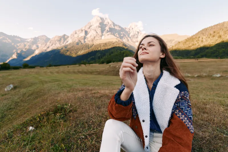 Woman portrait in meadow by mountains, nature outdoors scene showing a hiking break and relaxation, female traveler in warm jacket enjoying alpine landscape and clear sky.