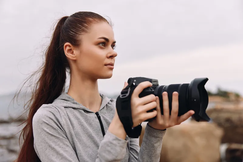 Young female photographer holding a camera near the beach, dressed in a gray hoodie, showcasing focus and creativity with a serene ocean backdrop