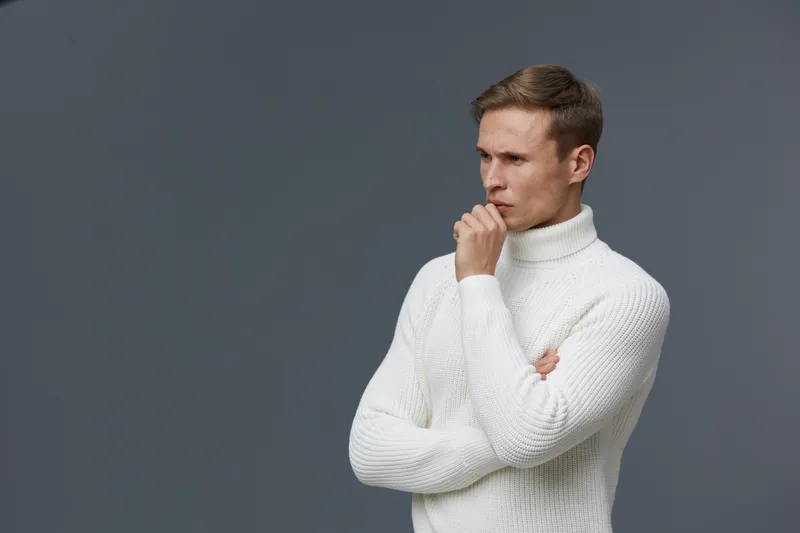 Thoughtful man wearing white turtleneck sweater isolated on gray background, pondering with hand on chin, contemplating or analyzing, vertical studio portrait, fashion and lifestyle concept.