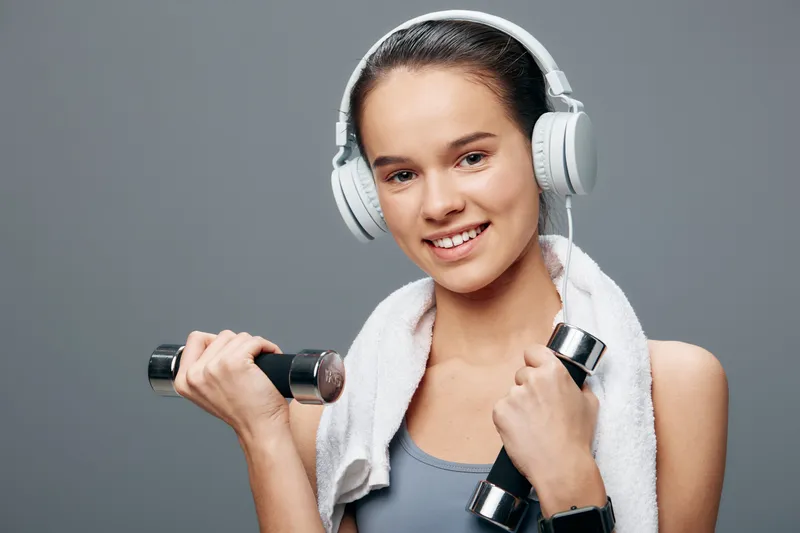 Woman fitness enthusiast in a gym, wearing headphones and a towel over her shoulders, gripping dumbbells and ready for a focused workout and training session.