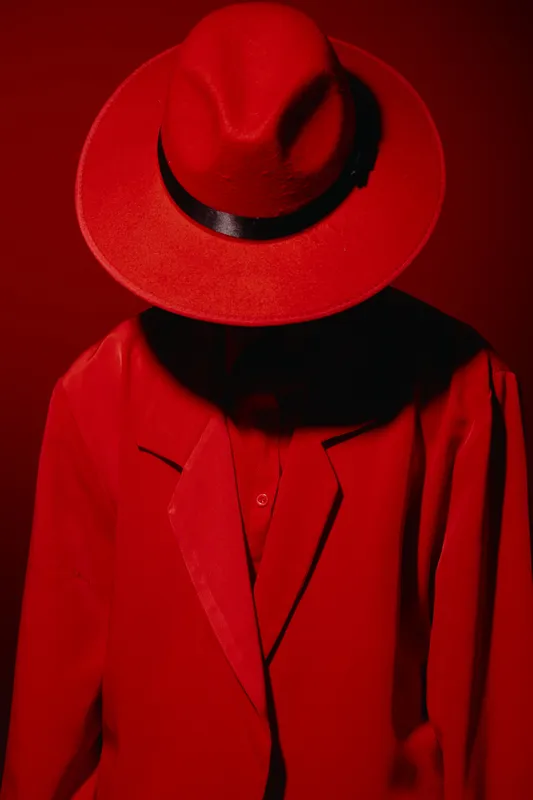 Fashion model wearing a bold red fedora hat and matching red jacket, obscuring the face with hat brim. Creative beauty concept with high contrast shadows on red background.