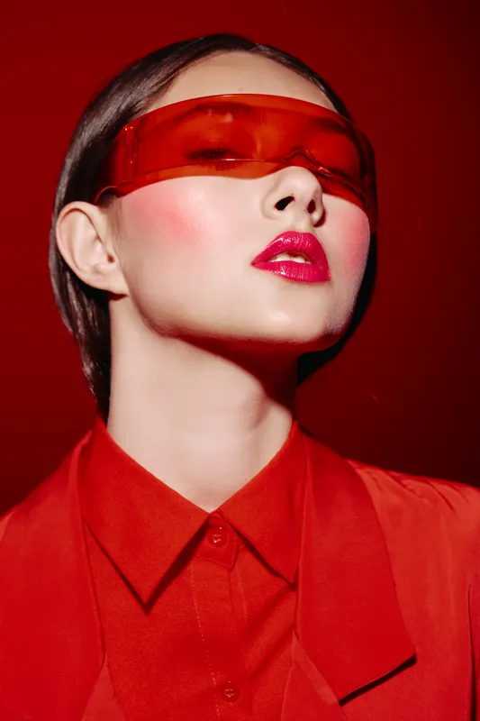 Fashion model with glossy bright pink lips and red sheer eyewear poses confidently in a red outfit. Creative beauty concept with bold colors and sharp light.