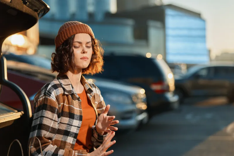 Woman, portrait, beanie, sunlight, cars, parking, serene, urban. Young woman in plaid jacket with eyes closed enjoying golden hour glow, authenticity and candid lifestyle vibe, mindful living and