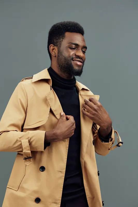 Stylish young man smiling while wearing a beige trench coat over a black turtleneck, set against a muted gray background, exuding confidence and fashion sense