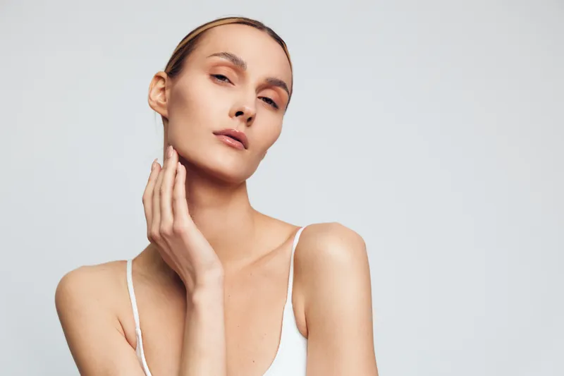 Portrait of a woman with clear skin and natural makeup touching her face, wearing a white top on a light gray background. Concept of beauty, skincare, and elegance.