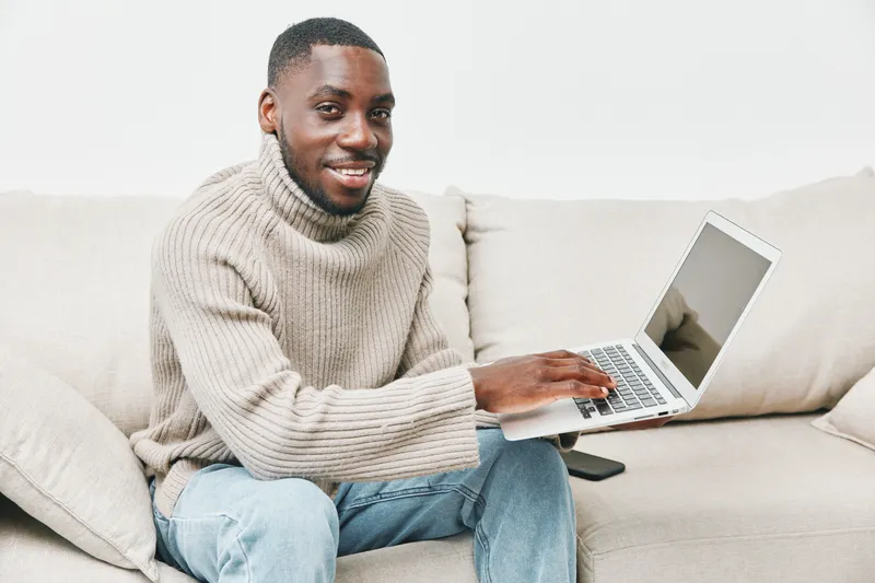 Smiling African American man working on a laptop at home in cozy sweater, showcasing remote work lifestyle and modern comfort