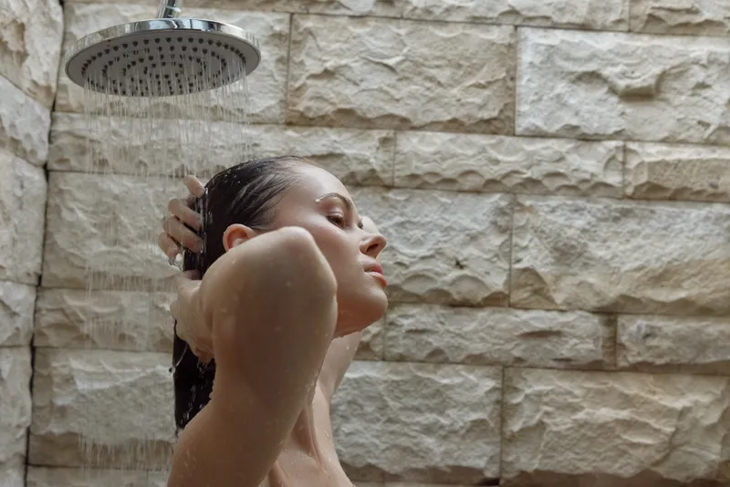 Woman taking shower under rainfall showerhead with natural stone wall background, relaxed expression, wet hair, indoor bathroom, soothing moment.