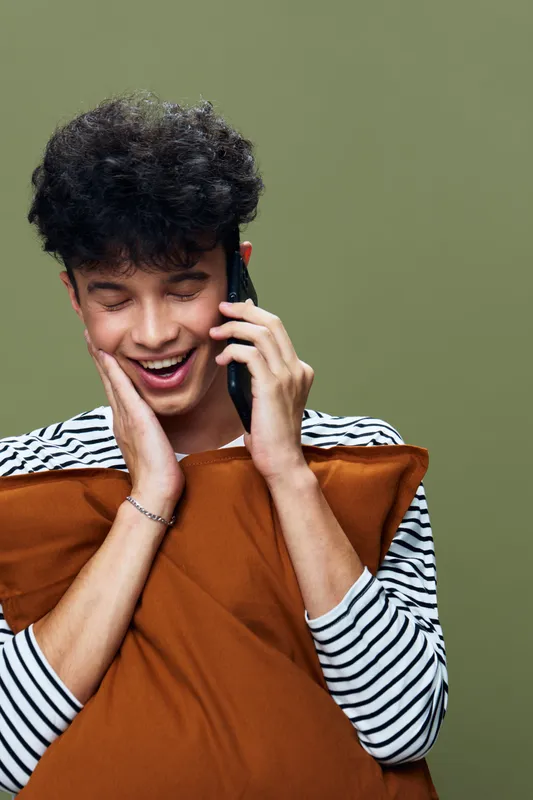Young man talking on phone with joyful expression, holding a brown pillow, wearing striped shirt against green background, casual indoor setting, cheerful mood.