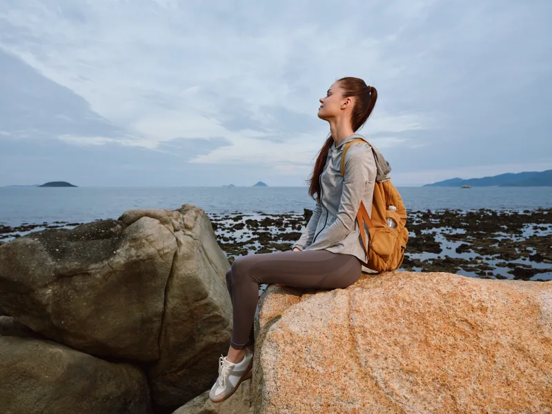 Young woman in athletic wear enjoying nature, sitting on a rock by the sea with a backpack, showcasing a relaxed and peaceful expression during sunset
