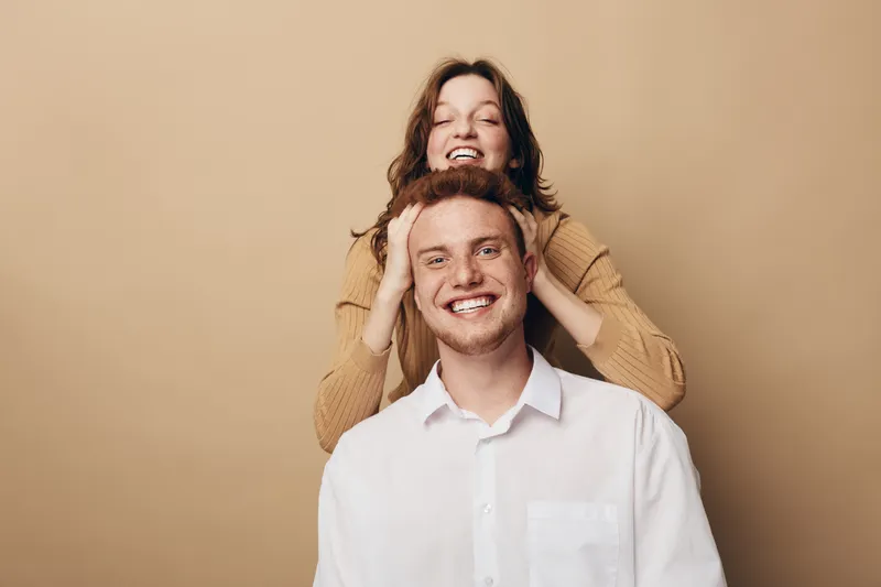 Happy young couple smiling and posing together on beige background. Woman stands behind man with hands on his head. Concept of friendship, joy, and positive emotions.