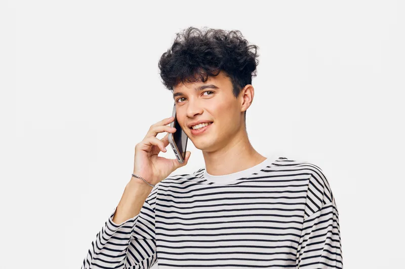 young man talking on smartphone wearing striped shirt, smiling and relaxed, isolated white background, casual communication concept