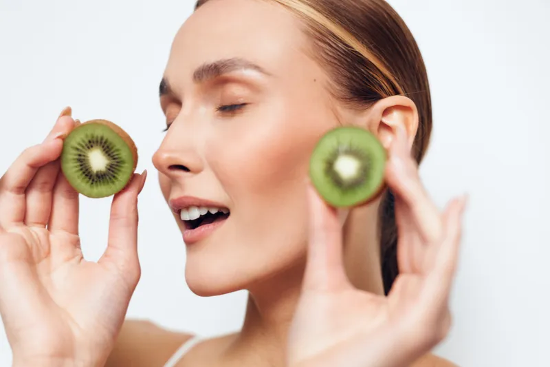 Young woman holding two kiwi halves close to her face, eyes closed, smiling gently, fresh fruit, healthy lifestyle, clean white background, natural beauty, wellness concept