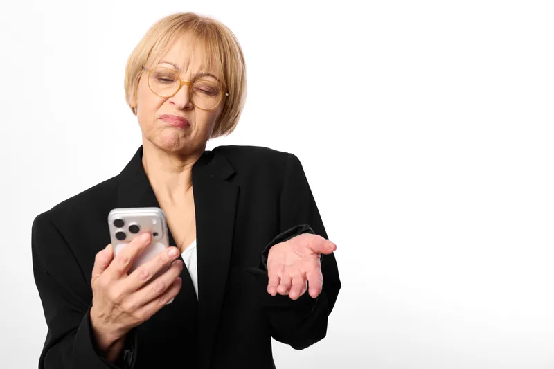Elderly woman in business suit looks stressed holding a smartphone, pointing forward with worried eyes, isolated on white background, communication concern