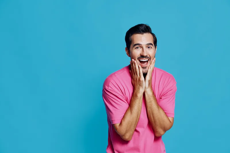 Handsome smiling man in a bright pink t-shirt expresses joy and excitement with his hands on his cheeks against a vivid blue background. Cheerful lifestyle concept.