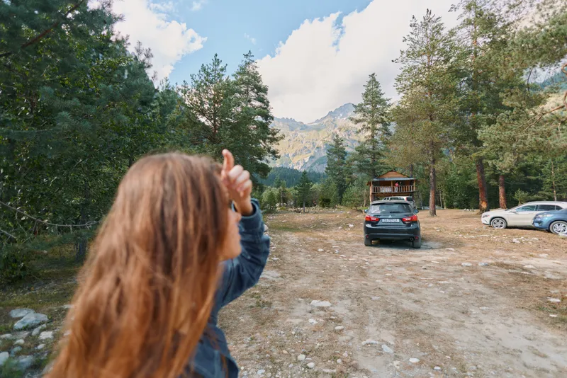 A lone traveler with long hair raises a hand toward distant mountains in a rugged forest setting, capturing a moment of exploration and awe.