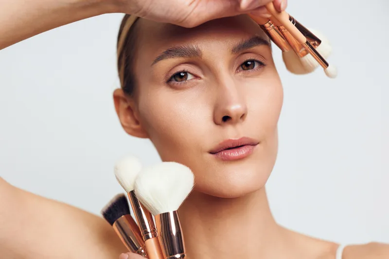 Close-up portrait of a woman applying makeup with brushes on her face, showing natural skin and beauty. Concept of cosmetics, skincare, self-care, and personal grooming in soft lighting.