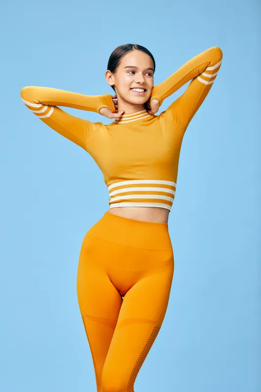Woman fitness model in bright orange athleisure posing with hands behind her head against a blue studio backdrop, showcasing confidence, flexibility, and athletic style for sport and wellness.