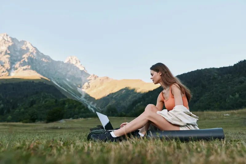 A young woman sits on a rolling mat in a grassy field with a laptop, mountains looming in the distance, creating a calm, productive outdoor work atmosphere.