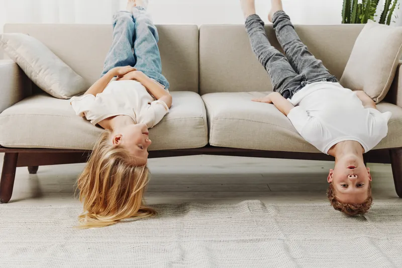 Two playful children lying upside down on sofa in casual clothes, enjoying relaxed moment at home with bright interior background.