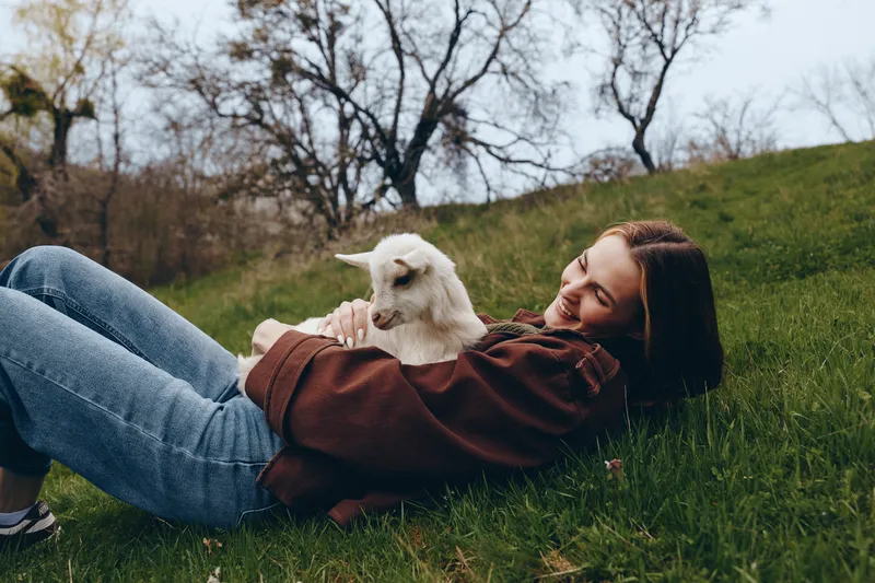 A young woman in a brown jacket lies on a grassy hill holding a white baby goat while smiling peacefully in a natural outdoor setting during early spring.