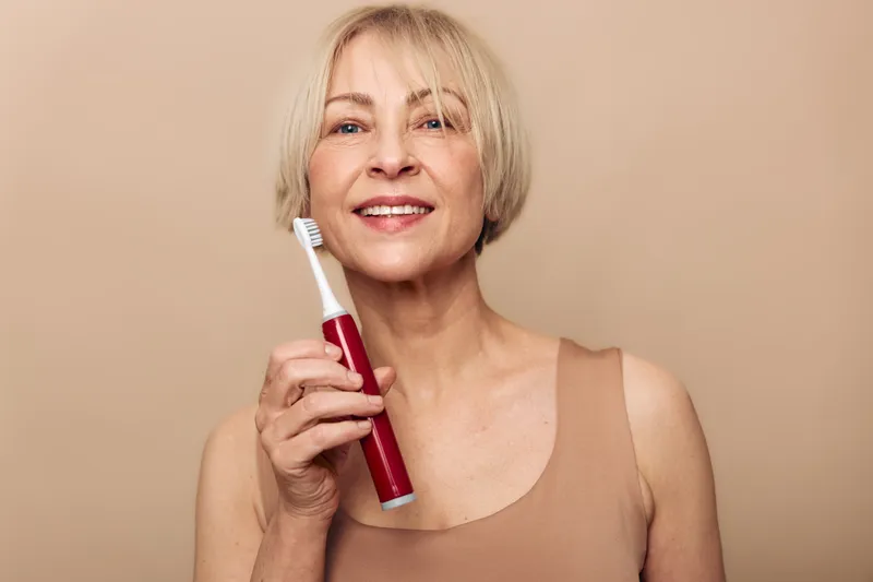 Smiling mature woman holding electric toothbrush isolated on beige background. Concept of dental care, oral hygiene, healthy lifestyle and senior wellness.