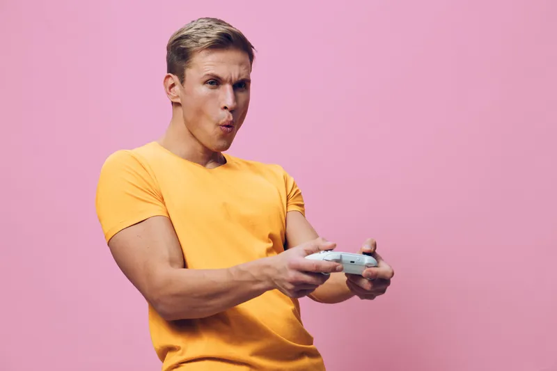 Smiling young man in yellow t-shirt holding game controller playfully looking at screen studio shot on pink background. Casual lifestyle concept