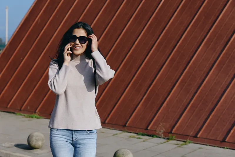 Woman in a light sweater and jeans talks on a smartphone outdoors, wearing sunglasses, with an orange slanted wall and concrete bollards, capturing casual urban communication.