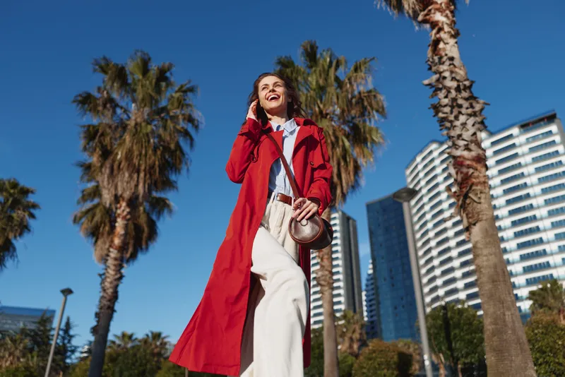 A stylish woman walks under clear blue skies in a bright red trench, posing against palm trees and modern high rises, exuding confident city fashion and urban chic.