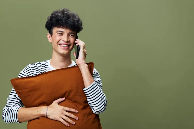 Young man smiling and talking on phone while hugging a brown pillow against green background, wearing a striped sweater and showing positive emotions.